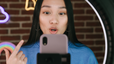 Image is a photograph of a young woman with black hair, using a selfie light and filming herself talking to the camera. Behind her is an interior brick wall with neon lights.