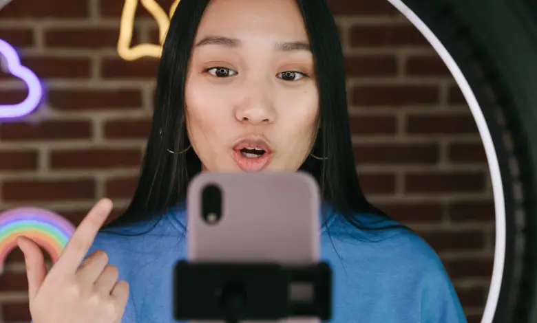 Image is a photograph of a young woman with black hair, using a selfie light and filming herself talking to the camera. Behind her is an interior brick wall with neon lights.