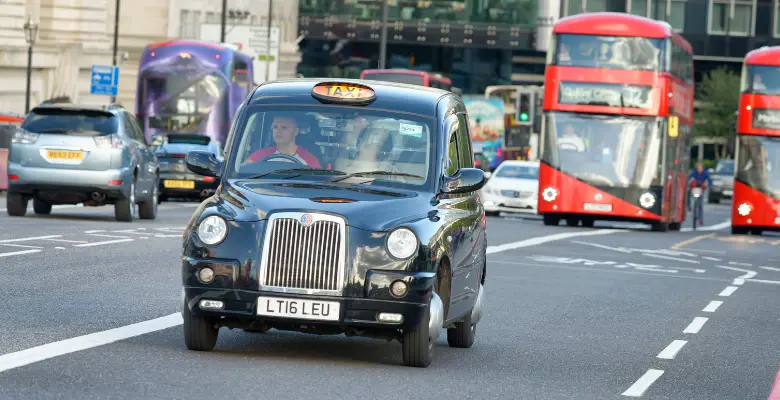 A black cab in London on a road with a red London bus behind it