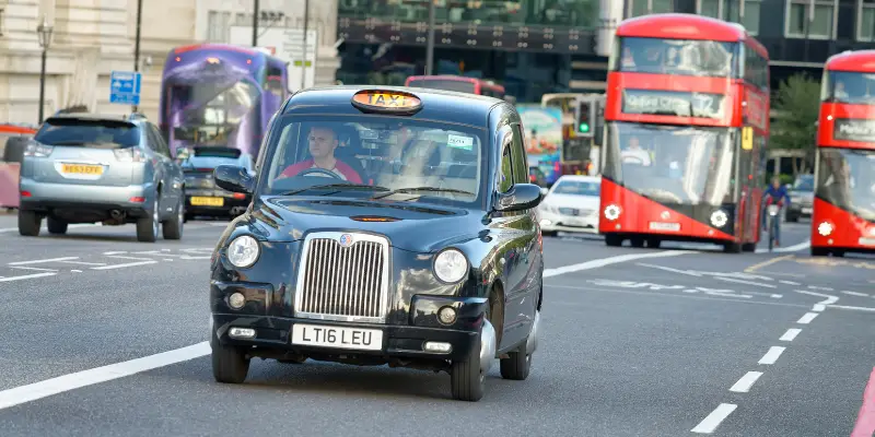 A black cab in London on a road with a red London bus behind it