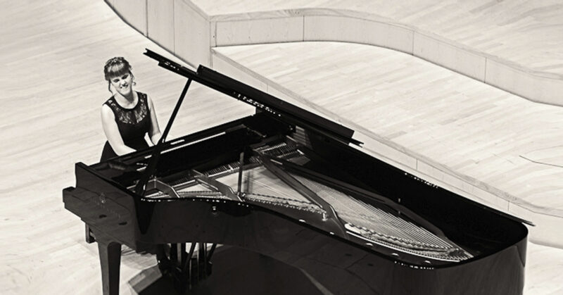 Image is a black and white photograph of Rachel Starritt playing a grand piano 