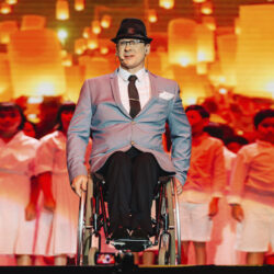 Tony Dee wearing a suit and hat sat in a wheelchair on stage, with a choir in the background
