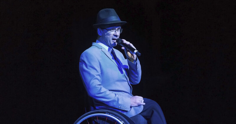 A photograph of Tony Dee singing on stage whilst in a wheelchair and with microphone in hand