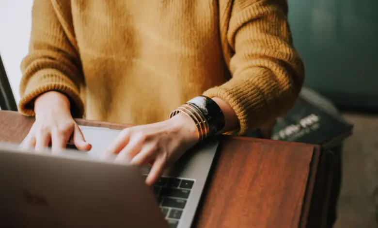 Photograph of a woman wearing a mustard-colour jumper and silver bangles, typing on a laptop on a wooden desk