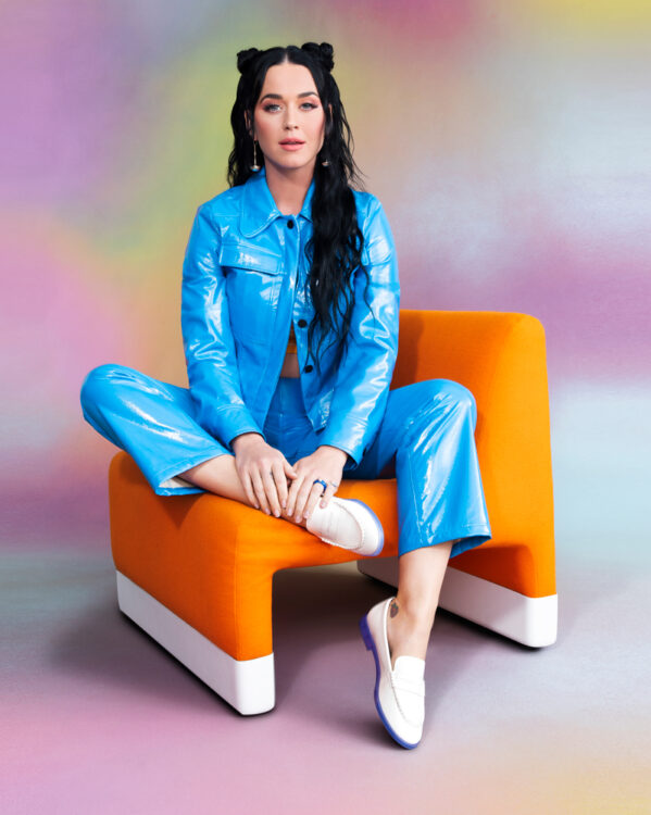 Photograph of Katy Perry sat on an orange chair wearing a bright blue suit, against a background of rainbow colours