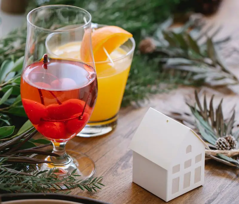 Two cocktails - one yellow with orange peel on the side and one red with berries in it - on a wooden table surrounded by foliage and a small white model of a house