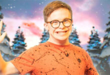 George Webster wearing an orange top and brown round glasses smiling and waving at the camera. He is stood in front of four Christmas trees.