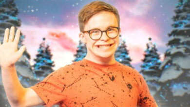 George Webster wearing an orange top and brown round glasses smiling and waving at the camera. He is stood in front of four Christmas trees.