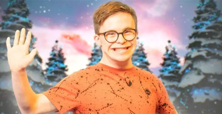 George Webster wearing an orange top and brown round glasses smiling and waving at the camera. He is stood in front of four Christmas trees.