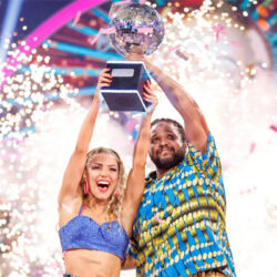Hamza Yassin and Jowita Przystal lifting the Strictly glitterball. They are wearing brightly coloured blue and yellow outfits that they performed in for their Couple's Choice, an Afrobeats-inspired dance. There are fireworks behind them.