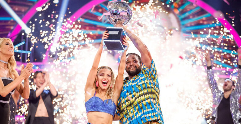 Hamza Yassin and Jowita Przystal lifting the Strictly glitterball. They are wearing brightly coloured blue and yellow outfits that they performed in for their Couple's Choice, an Afrobeats-inspired dance. There are fireworks behind them.