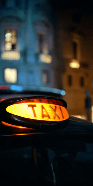 A close up of a taxi sign on top of a black cab at night. There is a building in the background. The words in red at the top say 'Have your say on possible increases to taxi fares in London, and words at the bottom in blue say 'Your views could change the outcome'.