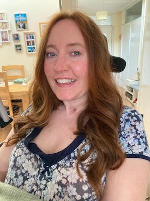 This photo shows Rebecca sitting in her wheelchair at home. It is a selfie she has taken and she is slightly to the right of the camera as you look at it. Rebecca has red hair, blue eyes and a wide smile.