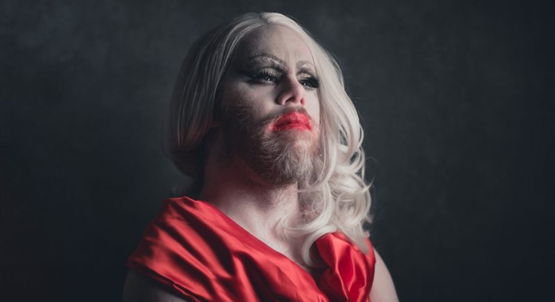Frozita Honkong with long platinum blonde hair and a full beard is wearing heavy makeup, including dark eyeshadow and bright red lipstick. They are dressed in a bright red garment. The background is dark and plain, which highlights their dramatic appearance.