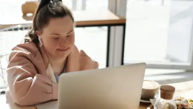 A woman with Down's syndrome sat in a cafe working at a table on her laptop. She has long brown hair in a ponytail and is wearing a soft pink jacket and pale blue top.