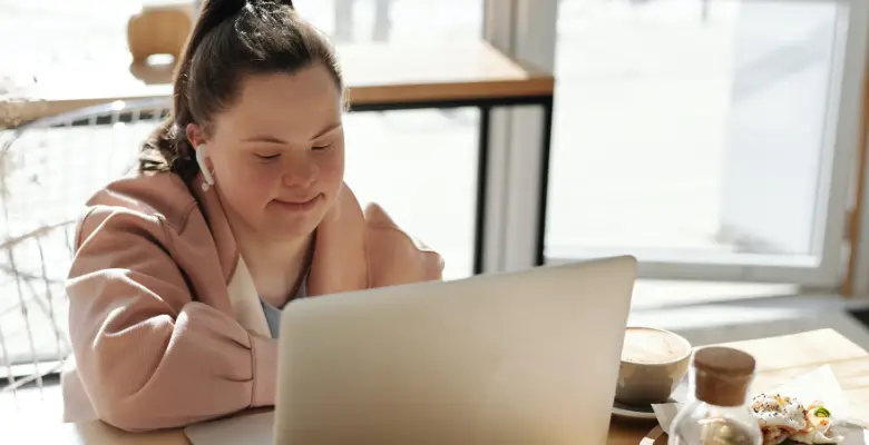 A woman with Down's syndrome sat in a cafe working at a table on her laptop. She has long brown hair in a ponytail and is wearing a soft pink jacket and pale blue top.