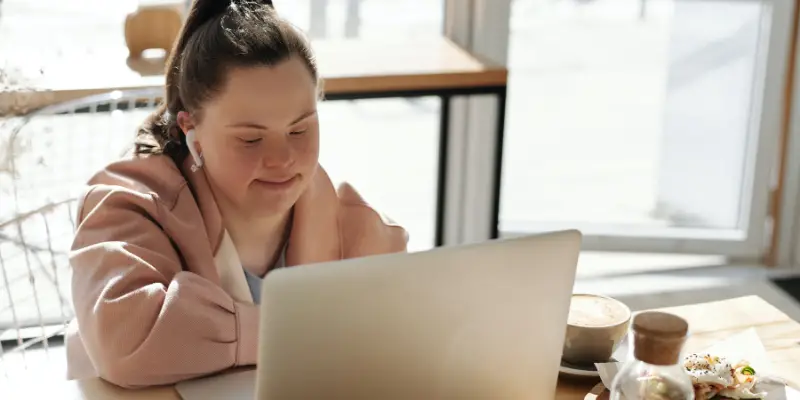A woman with Down's syndrome sat in a cafe working at a table on her laptop. She has long brown hair in a ponytail and is wearing a soft pink jacket and pale blue top.