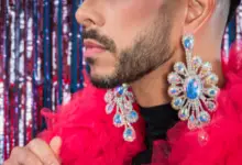 A close up of the lower part of a man's face. He has a black beard and has a pink feather boa around his neck and is wearing blue jewelled earrings