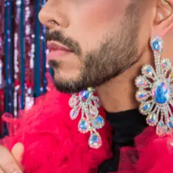 A close up of the lower part of a man's face. He has a black beard and has a pink feather boa around his neck and is wearing blue jewelled earrings