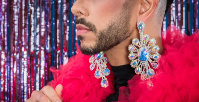 A close up of the lower part of a man's face. He has a black beard and has a pink feather boa around his neck and is wearing blue jewelled earrings