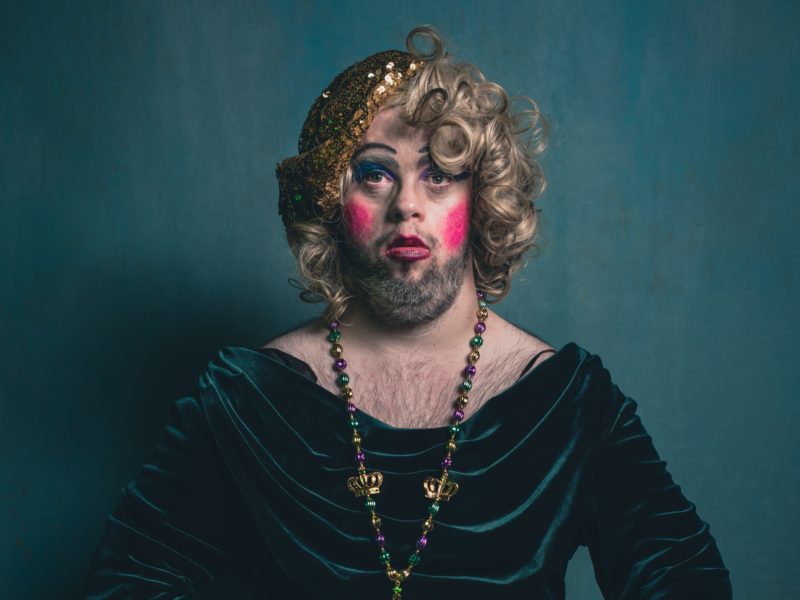 Horror Shebang from Drag Syndrome. A person with curly blonde hair and a full beard is wearing heavy makeup, including blue eyeshadow and bright pink blush. They are dressed in a dark green velvet top and a beaded necklace with purple, green, and gold beads, including small crown charms. The background is plain and dark, emphasizing the individual's expressive appearance.