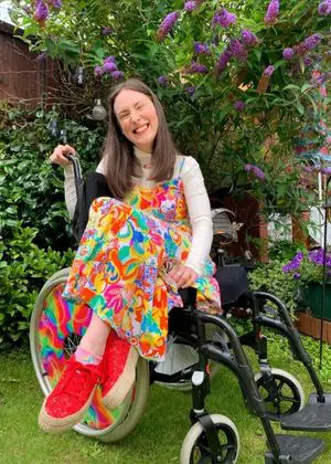 Rebecca sitting in her wheelchair with her legs out to the side. She is wearing a colourful printed dress with a log sleeve top underneath and red trainers with short colourful socks