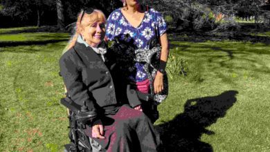 Clare, a wheelchair user, and her friend Sue stand together in a sunny park, smiling; they have been friends for over 30 years.