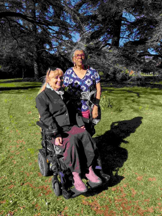 Clare, a wheelchair user, and her friend Sue stand together in a sunny park, smiling; they have been friends for over 30 years.