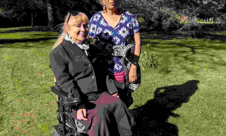 Clare, a wheelchair user, and her friend Sue stand together in a sunny park, smiling; they have been friends for over 30 years.