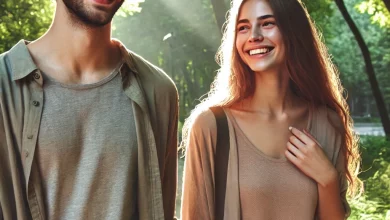 A young man and woman walking outdoors in a sunlit park, smiling warmly at each other. The man has short, wavy dark hair and wears a beige shirt over a gray t-shirt. The woman has long, wavy brown hair and wears a light brown top with a cardigan and jeans. Tall green trees and soft sunlight create a serene background.