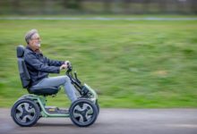 An older man with gray hair and glasses is riding a four-wheeled mobility scooter on a paved path. He is seated upright and smiling while holding the handlebars. The scooter has large tires and a supportive backrest, designed for outdoor use. The background shows a grassy field in motion blur, indicating movement.