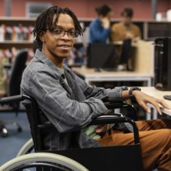 Young man in a wheelchair sitting at a computer desk in a library, smiling at the camera.