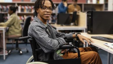 Young man in a wheelchair sitting at a computer desk in a library, smiling at the camera.