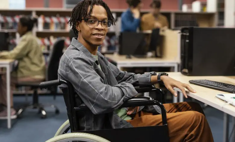 Young man in a wheelchair sitting at a computer desk in a library, smiling at the camera.
