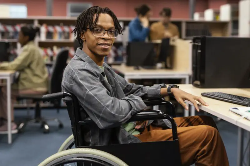Young man in a wheelchair sitting at a computer desk in a library, smiling at the camera.