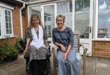 Two women sit outside a brick and glass house. One is in a power wheelchair and smiling, the other sits beside her on a wooden chair. The setting appears relaxed and friendly.