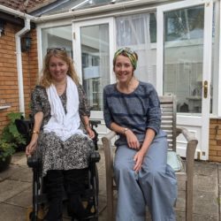 Two women sit outside a brick and glass house. One is in a power wheelchair and smiling, the other sits beside her on a wooden chair. The setting appears relaxed and friendly.