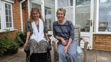 Two women sit outside a brick and glass house. One is in a power wheelchair and smiling, the other sits beside her on a wooden chair. The setting appears relaxed and friendly.
