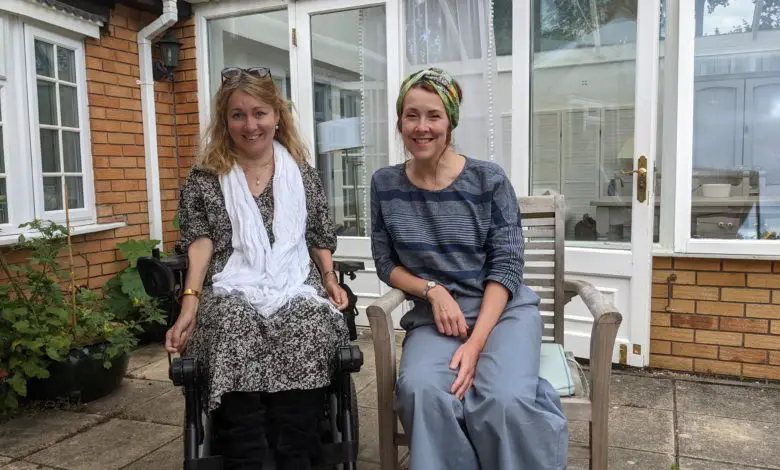 Two women sit outside a brick and glass house. One is in a power wheelchair and smiling, the other sits beside her on a wooden chair. The setting appears relaxed and friendly.