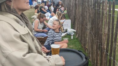 A woman in a power wheelchair watches an outdoor event from behind a fence, holding a beer on her tray. People sit on the grass beside her, facing the same direction. A crowd and scoreboard are visible in the distance under a cloudy sky.