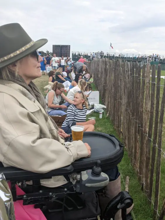 A woman in a power wheelchair watches an outdoor event from behind a fence, holding a beer on her tray. People sit on the grass beside her, facing the same direction. A crowd and scoreboard are visible in the distance under a cloudy sky.