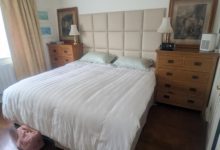 A double bed with a white duvet and green pillows, flanked by wooden bedside cabinets. A CPAP machine sits on the right cabinet next to a lamp and framed artwork.