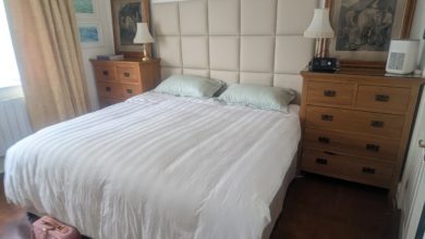 A double bed with a white duvet and green pillows, flanked by wooden bedside cabinets. A CPAP machine sits on the right cabinet next to a lamp and framed artwork.