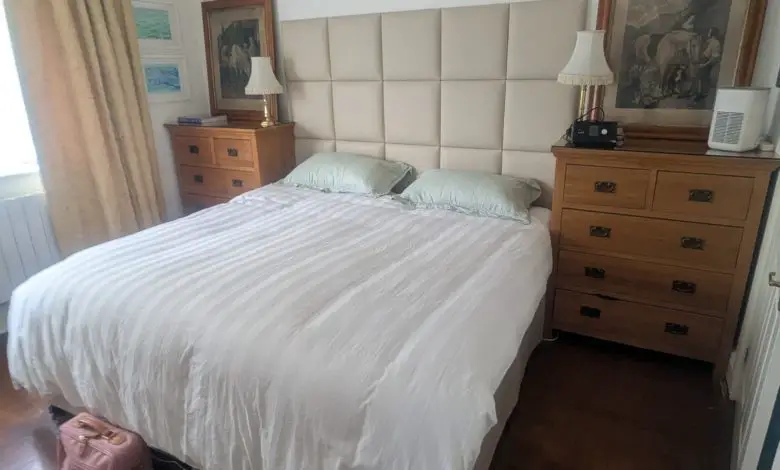A double bed with a white duvet and green pillows, flanked by wooden bedside cabinets. A CPAP machine sits on the right cabinet next to a lamp and framed artwork.