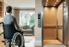 A man in a wheelchair faces an open home lift in a bright, modern living room with wooden flooring and a grey sofa near a window.