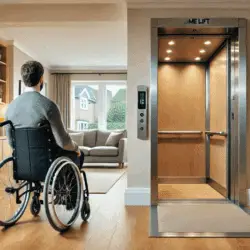 A man in a wheelchair faces an open home lift in a bright, modern living room with wooden flooring and a grey sofa near a window.