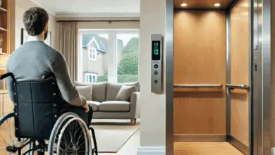 A man in a wheelchair faces an open home lift in a bright, modern living room with wooden flooring and a grey sofa near a window.