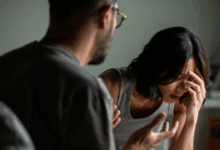 A distressed woman sits with her head in her hand while a man speaks to her in an animated, confrontational manner.