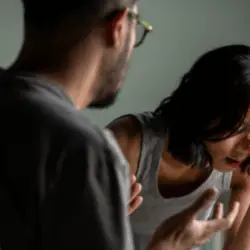 A distressed woman sits with her head in her hand while a man speaks to her in an animated, confrontational manner.