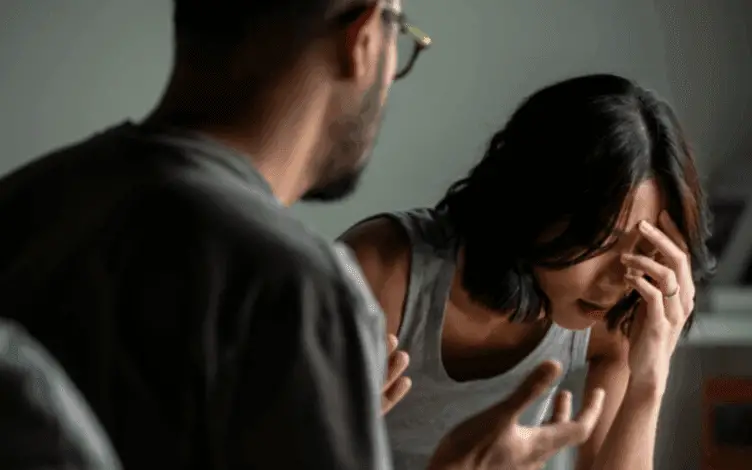 A distressed woman sits with her head in her hand while a man speaks to her in an animated, confrontational manner.